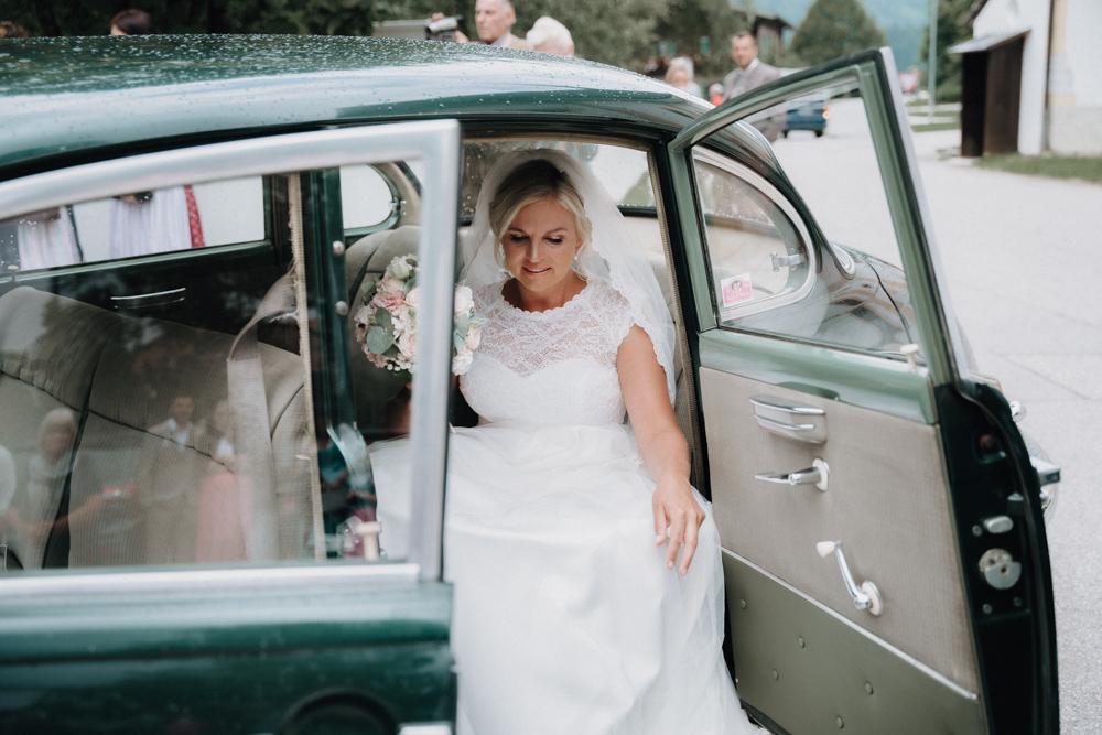 Braut steigt aus einem Oldtimer vor der Kirche in Inzell – Hochzeit im Pitzlloch beginnt.
